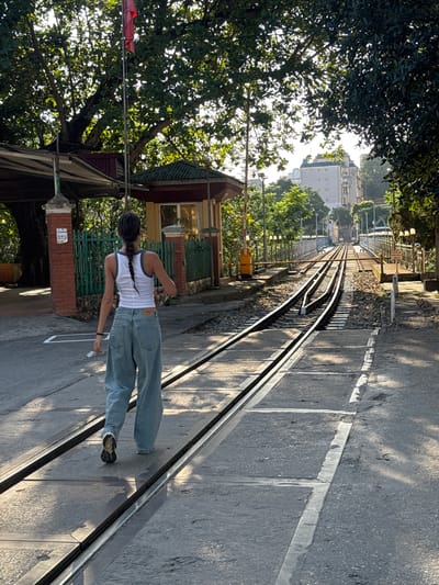 The image depicts a person standing on a narrow gauge railway track in Lao Cai Ward, Vietnam. The person, seen from the rear, is wearing a white tank top, loose blue jeans, and dark shoes. They have long dark hair styled in a braid. The person is situated on the tracks, which run parallel to a paved road. To the left, there is a verdant tree canopy and a building with a green roof. In the background, there are additional buildings and trees visible in the distance. The photograph is taken during daylight hours, as evidenced by the shadows cast on the road and railway tracks.