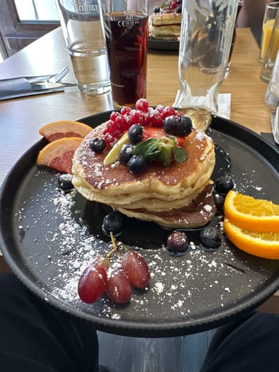 The scene depicts a breakfast or brunch meal served at a restaurant or cafe in Weißenburg in Bayern, Germany. In the foreground, a black, round plate prominently features a stack of three golden-brown pancakes, lightly drizzled with dark syrup or sauce and dusted with powdered sugar. The pancakes are topped with a cluster of fresh fruits including red currants, blueberries, and sliced strawberries, accompanied by a small green leaf. Surrounding the pancakes on the plate are three thin slices of pink grapefruit on the left, two orange wedges on the right, and a small cluster of three red grapes with a stem towards the bottom left. Additional individual blueberries are scattered on the plate, also dusted with powdered sugar.
On the light brown wooden table, to the upper left of the main plate, a clear glass of water stands beside a taller clear glass containing a dark beverage, marked "SOHM," "0,2l," and "CE M 23." Further back, a blurry clear glass, possibly empty, is visible. A fork and knife, wrapped in a dark napkin, are placed to the left of the water glass. In the blurred background, another black plate with a stack of pancakes topped with fruit is visible, indicating another diner at the table. A light-colored napkin or small paper is visible near the center-right. The shot is taken from a seated individual's perspective, with dark-colored pants visible at the bottom of the frame. The overall lighting suggests an indoor daytime setting.