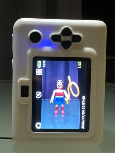 The image shows the back of a white handheld digital camera or gaming device. On the screen, a small figurine of Wonder Woman is visible, holding a golden lasso. The device has several buttons on the top, including a directional pad and what appears to be a circular button that is illuminated by a blue light. The screen displays a battery icon, a resolution of "12M", a zoom level of "1.0X", and a timestamp "2026/07/24 10:57:43". The device is placed on a reflective surface, possibly a table, with a dark background. There is no indication of the location being Fañabé, Spain.