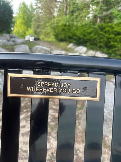 The image shows a close-up of a black metal bench with a bronze plaque that reads "SPREAD JOY WHEREVER YOU GO". The plaque is attached to the bench with screws. The background is blurred but appears to be a natural outdoor setting with trees, rocks, and a dirt path. A vehicle is faintly visible in the distance. The image is taken from a slightly downward angle, focusing on the plaque and the bench. The location is identified as Harbor Springs, United States of America.
