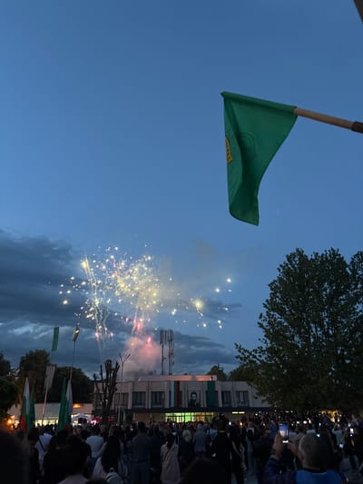 The image captures a vibrant public celebration at dusk or early night in Skravena, Bulgaria, characterized by fireworks illuminating the twilight sky.

In the upper portion of the frame, a large green flag, attached to a wooden pole, is prominently displayed against the deep blue sky. It features a yellow emblem or text, partially visible and appearing to be in Cyrillic script. Below and to the right, brilliant fireworks are exploding, showering bright sparks of yellow and white, with some red trails, across the sky and clouds.

The lower and right sides of the image are filled with a large crowd of people, densely packed together, all looking towards the fireworks. Many individuals are holding up their smartphones, actively recording the spectacle.

In the background, behind the crowd, are several buildings and trees. One building, in particular, stands out with a large portrait of a man displayed on its facade, suggesting a historical or national figure relevant to the location. Cellular antennae are visible on the rooftops of the buildings. Among the crowd and nearer to the buildings, other smaller flags are visible, including additional green flags and some that display the white, green, and red stripes characteristic of the Bulgarian national flag. The scene conveys a festive and communal atmosphere.