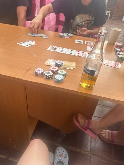 The image depicts a casual gathering at a wooden table, likely a card game, with chips and cards laid out. A partially filled bottle of beer with a label reading "Балтика" is prominently placed on the table. The setting appears to be outdoors, suggested by the wooden floorboards and natural lighting. In the foreground, a person's legs are visible, wearing pink slides and white Crocs. The overall atmosphere is relaxed and informal. There is no information in the image to suggest the location is in Zhebrei, Russia.