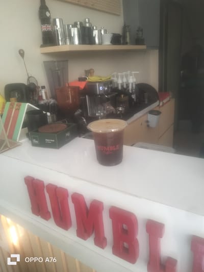 This media file depicts a coffee shop counter in Nusaherang, Indonesia. The foreground is dominated by a white counter with large, red, three-dimensional letters spelling out "HUMBLE" across the front. To the right, on the counter, sits a clear plastic cup filled with a dark beverage, likely iced coffee, with the "HUMBLE Coffee" logo visible in red text on the cup. Behind the cup, more of the counter is visible, lined with various coffee-making equipment, including a coffee machine and dispensers.
On a shelf above the counter, several objects are visible, including what appears to be a decorative replica of Big Ben, various metal shaker-like containers, and a white ceramic cup. The background is blurred, but suggests a working coffee shop environment. The lighting appears to be indoor and somewhat dim, possibly indicating an evening or overcast day. No people are visible in the frame. The overall impression is a clean and organized coffee service area, with the prominent "HUMBLE Coffee" branding suggesting the name of the establishment. The bottom left corner displays "OPPO A76," indicating the device used to capture the image.