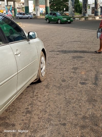 The image shows a street in Ado Ekiti, Nigeria. A silver Toyota Camry is parked on the right side of the street, facing the viewer. The car has its driver's side door open, suggesting that the driver has just exited the vehicle. A green car, possibly a Toyota Corolla, is driving in the distance, moving to the right of the frame. A woman in pink and black clothing walks away from the camera on the right side of the frame. The woman is holding a black bag. The street is paved with asphalt and has a lot of small, dark rocks on the surface. The environment is dusty, indicating a dry climate. A billboard is visible in the background on the left. A highway overpass is visible in the background. The image captures a typical moment on a street in a Nigerian city.