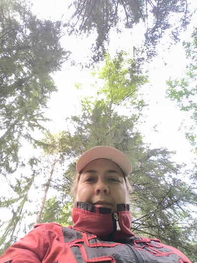 A low-angle, outdoor, waist-up selfie shows a woman looking directly at the camera from a forest. The woman is wearing a pink baseball cap and a red and gray jacket. The forest canopy with green leaves and branches fills the background, with bright sunlight filtering through. The scene is captured during the daytime. The location is unspecified, but it is claimed to be in the city of Voskhod 1, Russia.
