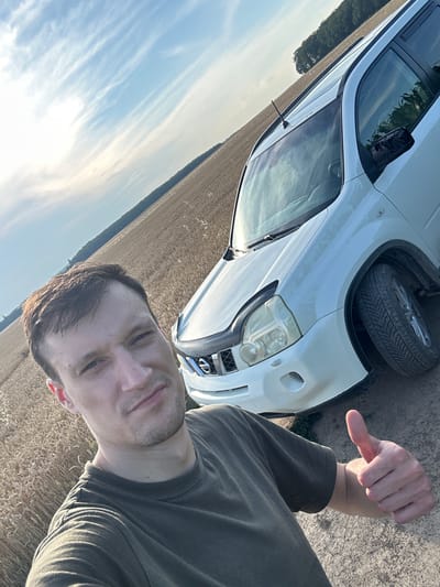 A low-angle, full-body selfie is taken by a Caucasian male with short brown hair and light stubble. He is wearing a dark olive-green t-shirt and is standing in a field of dry, tan wheat under a bright blue sky with wispy white clouds. To his right, a white SUV is parked, its front fender and tire visible. The man is giving a thumbs-up gesture with his right hand. The background consists of the wheat field extending to a line of trees in the distance. The scene appears to be in the countryside, possibly near Slobidka-Zalisetska, Ukraine, based on the provided context.