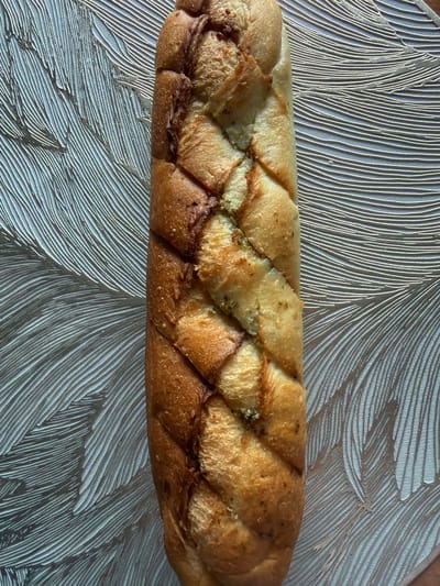 A single loaf of braided, herb-infused bread lies horizontally on a textured, silver surface that resembles flowing lines or feathers. The bread has a golden-brown crust with darker, toasted areas and visible green specks of herbs embedded in its crevices. The surface beneath the bread is highly reflective and appears to be a placemat or tablecloth with a repeating, linear pattern. The lighting casts subtle shadows, highlighting the texture of both the bread and the surface. There is no indication of the city of Kurgan, Russia, or any people, actions, or interactions in this image.