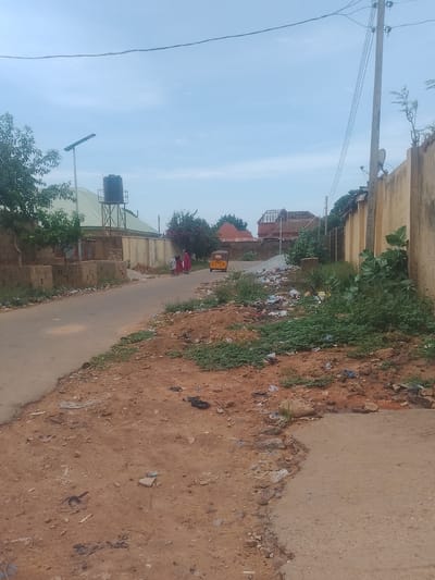 A street scene in Gindi Nassarawa Kyarmi Galbi, Nigeria, shows a paved road with scattered litter and vegetation along its edges. In the background, several buildings are visible, including one with a large water tank on a metal structure and another with a green roof. A solar-powered street light stands to the left. Three individuals wearing traditional clothing walk along the road on the left side. A yellow motorized tricycle is parked further down the road on the right. A tall concrete utility pole with wires runs along the right side of the image, adjacent to a beige-colored wall. The sky is partly cloudy and light blue.