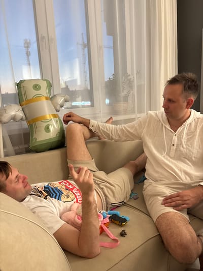 Two men are on a couch. The man on the left is lying down, looking up, with his head resting on a pillow. He is wearing a white t-shirt with graphics and beige shorts. The man on the right is sitting up, leaning back, with his left leg extended and his right leg bent. He is wearing a light-colored hooded shirt and beige shorts. A money-themed balloon is to the left of the men. Various small toys are scattered on the couch between them. In the background, through a window with white curtains, construction cranes and a cell tower are visible, suggesting an urban or suburban environment. The overall scene appears to be an indoor living space, possibly an apartment, in Красный Бор, Russia.