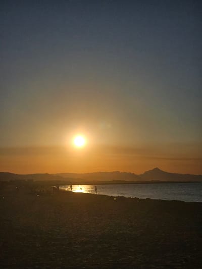 The image depicts a sunset over the beach in Deveses, Spain. The sun, a bright orb, is positioned in the center of the frame, casting a warm golden light across the sky. The horizon is marked by silhouetted mountains and the calm sea. Several people are visible on the beach and in the shallow water, some walking and others appearing to be engaged in recreational activities. The beach itself is composed of dark sand, with a few scattered lights suggesting some form of illumination or beach infrastructure. The overall scene conveys a serene evening at the seaside.