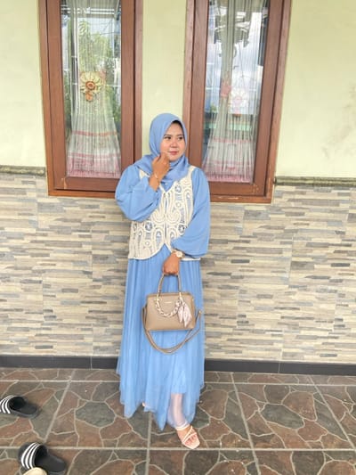 An adult female stands outdoors against a textured wall in Ketegan, Indonesia. She is positioned slightly right of center, looking towards the right side of the frame, and her left hand is raised to her chin. She wears a light blue hijab, a matching long-sleeved dress or tunic, and an off-white crocheted sleeveless vest. A light-colored watch is visible on her left wrist. In her right hand, she holds a light brown handbag adorned with a gold-colored chain on the handle and a patterned silk scarf. She is wearing light-colored open-toed sandals.
The background features a two-toned wall; the lower section consists of horizontally stacked, rectangular stone or tile pieces in varying shades of grey and brown, while the upper section is a plain light yellow. Two brown-framed windows with visible curtains and reflections of foliage are present in the upper background. The subject stands on a tiled floor with irregular, multi-toned grey and brown patterns. To the lower left, two pairs of slippers are visible on the ground: one pair of black and white striped slides and one light-colored slipper.