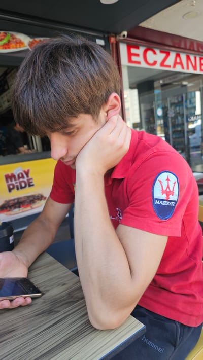 A young man with brown hair rests his head on his hand, looking down. He is wearing a red polo shirt with a Maserati logo on the sleeve and dark pants with the word "hummel" on them. He is sitting at a wooden table, holding a smartphone in his left hand. In the background, a sign for "ECZANE" (pharmacy) is visible on a building, along with a yellow sign advertising "MAY PIDE." The scene appears to be outdoors, likely in a public area in Ataşehir, Turkey, possibly a cafe or restaurant.