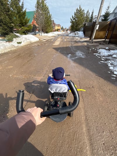 This image captures a first-person perspective of an individual pushing a child on a tricycle along an unpaved, muddy residential street in Gamovo, Russia. In the immediate foreground, a person's right arm, covered by a brown sleeve, grips the black, textured handle of a child's tricycle. A small child, wearing a blue jacket and a dark blue beret-style hat, is seated on the tricycle's grey seat, facing away from the camera and holding the tricycle's handlebars. A yellow object, likely a toy, is visible extending from the back of the tricycle.
The road surface is wet, composed of dark brown, compacted dirt and gravel, showing visible tire tracks and numerous small puddles reflecting the sky. Melting snow patches are present along the sides of the road. To the left, a low wall fronts a property with evergreen trees, behind which a house featuring a green roof and red upper facade is partially visible. Further along the street on both sides, various residential houses, fences, and gates are discernible, interspersed with more evergreen and bare deciduous trees. A utility pole stands on the right side of the road. The sky is a light blue with some scattered white clouds, and shadows cast across the road suggest sunlight from a high angle.