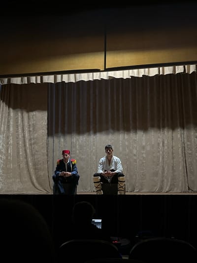 The image depicts a stage, possibly in Voiniagovo, Bulgaria. Two actors are present. One is wearing a white shirt and appears to be restrained with chains around their wrists. The other is wearing a dark-colored suit with a red fez. They stand in front of a beige curtain that serves as the backdrop. A dark-colored partition or screen is visible on the left side of the stage, and dark stage equipment is visible on the right. The lighting is dim and uneven.