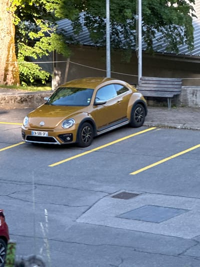 A golden Volkswagen Beetle is parked in a parking lot marked with yellow lines. The car has a license plate that reads "EN 586 OF". In the background, there is a wooden bench and some trees. The scene is likely in an outdoor urban or suburban setting in Le Sentier, Switzerland.