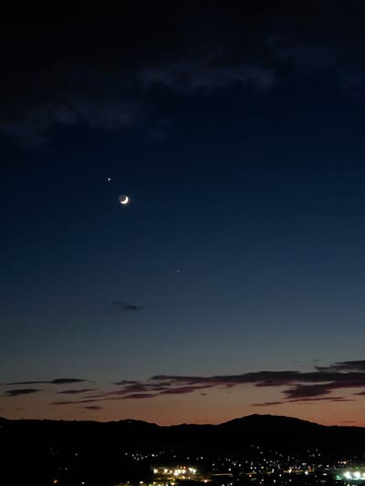 An astronomical scene captured at twilight above the Municipal Unit of Aegio, Greece. The image features a crescent moon and a bright planet in the dark blue sky. Below, a silhouetted mountain range frames a distant cityscape with scattered lights. The horizon is marked by a gradient of colors transitioning from deep blue to orange and pink hues, indicative of sunset. There are no people or animals visible in the frame. The overall environment suggests a serene, natural setting juxtaposed with urban development.