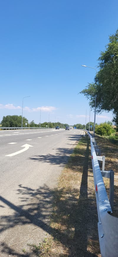 The image depicts a wide, paved highway with multiple lanes under a clear blue sky. On the left side of the road, a large white arrow is painted on the asphalt, indicating a turn. Several vehicles are visible on the highway in the distance. Along the right side of the road, a metal guardrail separates the highway from a grassy area with sparse vegetation and scattered dry grass. Tall, slender streetlights line the highway at regular intervals. The scene is bathed in bright sunlight, casting distinct shadows of tree branches and foliage onto the road and the grassy verge. The environment appears to be a rural or semi-rural setting, typical of a highway bypass. There are no visible people or buildings. The image appears to be taken from a low angle, close to the ground, facing the direction of traffic.