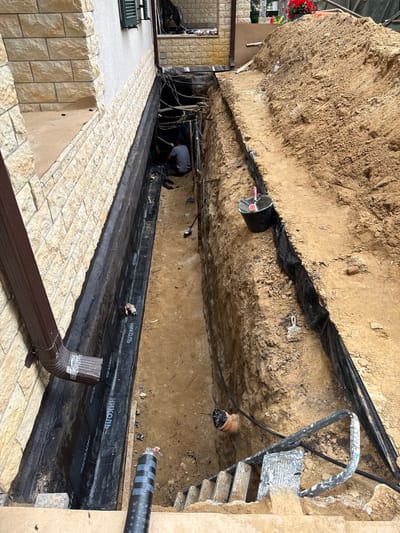 The image depicts an outdoor construction site in the city of Sosny, Russia. A deep trench has been excavated alongside the foundation of a building. The trench is lined with black waterproofing material, and the excavated earth is piled high on one side. A person wearing a grey t-shirt is visible working in the trench. Construction materials and tools, including a bucket, a shovel, and rolls of material, are scattered around the site. A downspout from the building extends down into the trench area.