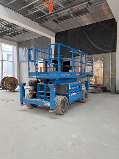 This image depicts an indoor construction or renovation site in Aulnay-sous-Bois, France. The primary subject is a blue, off-road scissor lift, positioned centrally and facing slightly left. The lift, branded "Acces," has its outriggers extended and features dirty, treaded tires. An identification number "11387" is visible on the rear of its chassis. No individuals are present on or near the equipment.

The environment consists of an unfinished interior space with a light gray, dusty concrete floor. The ceiling is exposed, revealing a grid of metal framework, pipes, and bundled orange electrical cables. The left wall features large window panes looking out onto an adjacent building with multiple stories. To the left, a large wooden cable spool with "5G16" and "Aulnay" spray-painted in red is visible. The rear and right walls are partially completed, displaying exposed concrete, sections covered with dark black and light gray tarps, and an opening on the right side obscured by translucent plastic sheeting. Pallets of materials are stacked in the background on the right, near a ladder-like metal structure against the wall. The overall scene suggests an active construction phase.