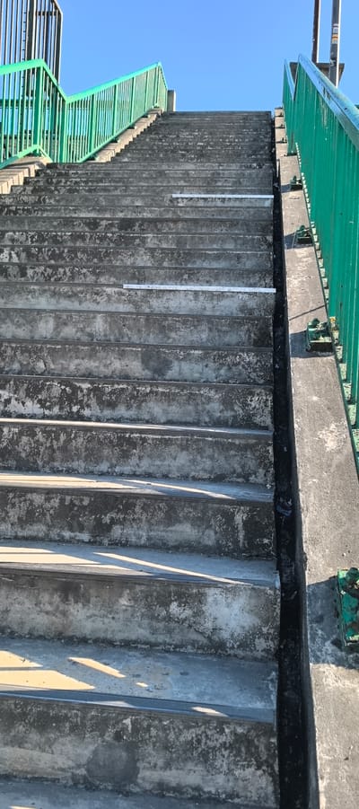 The image shows a concrete staircase with green railings on either side. The staircase is outdoors, under a clear blue sky. The concrete steps are worn and show signs of age and weathering. There are two horizontal white markers placed on the steps, one approximately one-third of the way up and the other about two-thirds of the way up. The railing on the left is higher and appears to lead to an elevated walkway or platform. The railing on the right is lower and runs along the side of the staircase. The staircase is located in Villeneuve-le-Roi, France, and appears to be part of a public walkway or bridge. There are no people or animals visible in the image. The environment is urban, with a clear view of the sky and no other structures or vegetation in the immediate vicinity of the staircase.