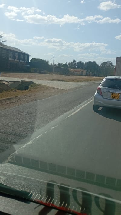 A silver Toyota Vitz with license plate KCT 647U is on a paved road, viewed from inside a vehicle with part of the dashboard visible. Beyond the road, a flat area with sparse vegetation leads to a building with dark windows. Another building is visible in the distance with a sign reading "H-WOOD DOORS". The sky is blue with scattered white clouds.