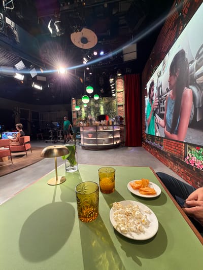 This image depicts a television studio set. In the foreground, a light green table holds two glasses filled with amber liquid, a small plate of potato chips, and a plate of popcorn. A brass lamp with a circular base is also on the table. In the midground, a bar area with shelves stocked with items is visible, along with a red curtain. Further back, studio lighting equipment is suspended from the ceiling. On the left side of the frame, two people are seated in orange chairs, and a camera operator is visible behind a camera. A large screen displays an image of two individuals talking. This television studio is located in the city of Sant Joan Despí, Spain.