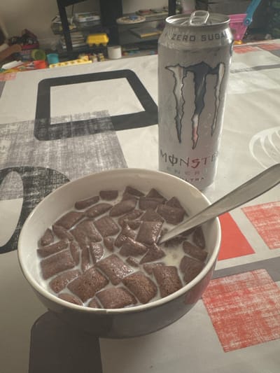 A can of Monster Energy Ultra Zero Sugar drink lies horizontally beside a bowl of chocolate cereal with milk and a spoon on a tabletop with a patterned tablecloth. The photograph appears to have been taken indoors. There is no information to connect this image to Welshpool, United Kingdom. No people are visible in the image.