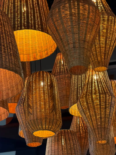A cluster of multiple illuminated, woven wicker or rattan lampshades hangs from a dark ceiling, filling the frame. The lampshades vary in size and shape, including bell-shaped, elongated conical, and cylindrical forms, all featuring a discernible woven texture. Each shade contains an internal light source, emitting a warm, orange-yellow glow that diffuses through the material and illuminates the open bottoms of several shades. Individual light bulbs are visible within some of the more transparent or open-ended shades. The overall impression is an interior decorative lighting arrangement. The scene is located in Fraccionamiento Los Heroes Queretaro, Mexico. No individuals or discernible activities are present.
