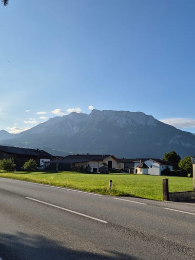 This vertical, eye-level, medium shot depicts a rural Austrian landscape under a clear blue sky. In the foreground, a paved road with a white dashed line runs horizontally across the frame, with its left side shaded by an unseen object. To the right of the road is a vibrant green grassy field. In the middle ground, several traditional Austrian farm buildings with dark roofs are visible, nestled amongst trees. A small, white chapel with a dark roof stands out among the buildings. In the background, a majestic mountain range dominates the scene, its peaks and slopes covered in a light haze. The overall impression is one of serene natural beauty and rural tranquility.