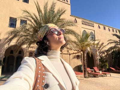 A single female individual is centrally featured in a sunlit outdoor scene located in Rissani, Morocco. She is presented in a selfie-style composition, visible from the chest up, wearing round sunglasses, a multi-colored patterned headscarf, a cream-colored ribbed turtleneck sweater, and a cream-colored jacket. A brown suede-like shoulder bag with silver studs rests on her left shoulder. Her head is tilted upward and slightly to the right, with her gaze directed towards the upper right of the frame.

The background consists of a large, multi-story building of light tan stucco or adobe construction, exhibiting traditional Moroccan architectural elements such as arched windows with intricate latticework, rectangular windows, and a crenellated roofline. Several tall palm trees are interspersed between the foreground and the building, casting distinct shadows. The ground in the mid-ground is paved with light-colored bricks or tiles. To the right, outdoor furniture includes multiple red-orange lounge chairs with wooden bases and a small table. A wooden pergola structure is also visible. The sky is clear and blue, indicating strong, direct sunlight.