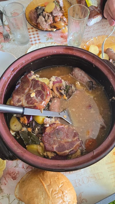 This is a top-down view of a meal setting. A large, brown ceramic pot dominates the center, filled with what appears to be a stew or casserole. The stew contains large pieces of meat, sausages, potatoes, and other ingredients in a broth-like liquid. A metal serving spoon is partially submerged within the pot.

To the left of the pot, on a floral tablecloth, there is a plate with a serving of the stew, two empty drinking glasses, and silverware. There's also packaged food. To the right of the pot, there is a loaf of bread.

Based on other sources, it can be inferred that this image was taken in Susam, Bulgaria. However, this cannot be determined from the image alone.