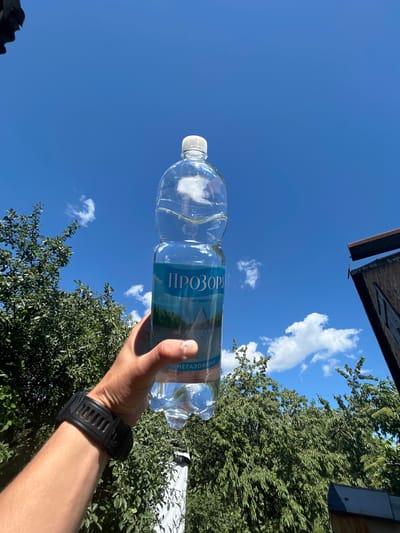 In a bright, sunny outdoor scene, a hand wearing a black digital watch holds a clear plastic bottle of "Prozora" brand mineral water. The bottle, filled with water, is positioned in the foreground against a backdrop of a vibrant blue sky dotted with white clouds. Lush green trees are visible on the left and right sides of the frame. On the far right, a portion of a rustic, weathered building with a metal roof is present. The image appears to be taken in Velykyi Trostianets, Ukraine, as indicated by the bottle's label.