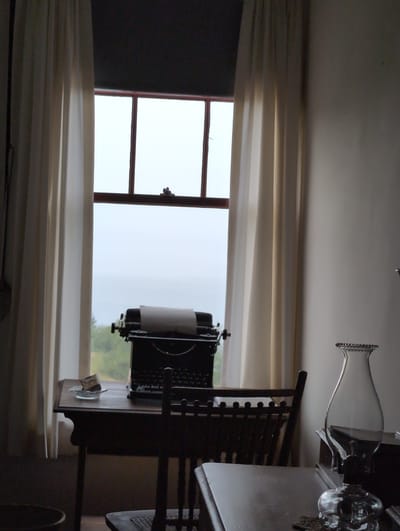 The image depicts an interior scene with a large window framed by off-white curtains. The window looks out onto a foggy or overcast sky and a hint of greenery, suggesting a natural landscape. In the foreground, a dark wooden desk sits with a vintage typewriter on it, a sheet of paper in its carriage. To the left of the typewriter, on a small glass dish, is a small packet of what appears to be tobacco or snuff. A wooden chair with a woven seat is positioned in front of the desk. To the right of the desk, on a dark surface, is a clear glass oil lamp with a decorative rim. The overall impression is of a rustic or historical setting, possibly a study or office. The location is identified as Campobello Parish, Canada, which is a rural area known for its natural beauty and historical significance. There are no people or animals visible in the image.