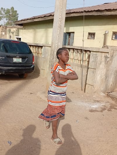 An outdoor scene in Gayawa, Nigeria, depicts a young person standing on an unpaved, reddish-brown dirt surface under a clear sky. The individual, identified as a girl with braided hair, wears a horizontally striped short-sleeved top (orange, white, blue) and a patterned reddish-brown skirt with blue accents, along with light-colored sandals. She stands with her arms crossed, facing slightly right. Her shadow is cast on the ground to her left.

To the left, a black Ford Focus station wagon is parked, facing away from the viewer, displaying a Nigerian license plate "KANO GADU." In the background, a light green single-story building with a corrugated metal roof and multiple windows, one featuring a blue shutter, is visible behind a concrete balustrade. A tall, grey concrete utility pole stands to the right of the person, with a metallic rod attached and extending downwards. Another building is partially visible on the far left, and a tree with green foliage is present in the upper left background. The environment appears dusty, indicative of an unpaved urban or semi-urban area, illuminated by sunlight.