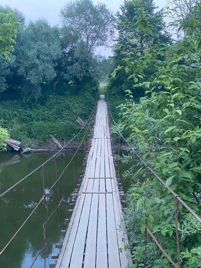 The image depicts a rustic, wooden suspension bridge spanning a calm, green river. The bridge is constructed with white-painted wooden planks for the walkway and reinforced with metal cables and posts. Lush, green foliage lines both banks of the river, with trees forming a dense canopy overhead. The sky is overcast and a pale gray, suggesting either early morning or a cloudy day. The scene is tranquil and natural, hinting at a rural or semi-rural setting. It is located in the city of Yuryatino, Russia.