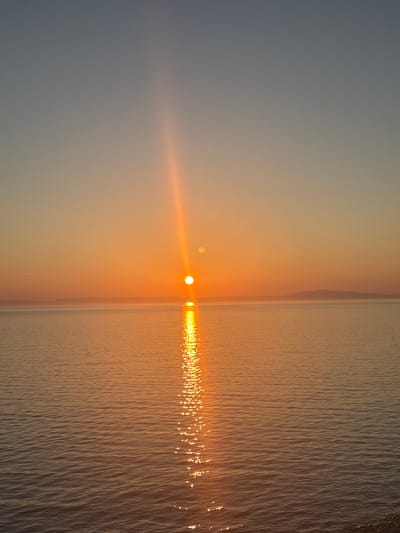 The image depicts a tranquil sunset over a body of water. The sun, a brilliant orb of orange, is positioned near the horizon, casting a radiant glow across the scene. A luminous reflection of the sun shimmers on the water's surface, creating a path of light. In the distance, a silhouette of hills or mountains can be seen against the warm hues of the sky. The sky itself transitions from a soft blue at the top to a warm orange near the horizon, a typical characteristic of a sunset. The water appears calm, with gentle ripples disturbing its surface. The foreground shows a dark, rocky embankment. This scene is located in the city of Turka, Russia.