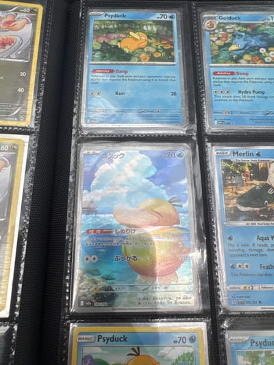 The image shows a close-up, eye-level view of a black binder filled with Pokemon cards. The binder is oriented vertically. Several Pokemon cards are visible within the plastic sleeves of the binder. On the top row, from left to right, are parts of three cards: the first is largely obscured but shows the top of a yellow border and the number "HP 60", the second card features a bright blue background with an orange Pokemon character and the text "Psyduck" and "70 HP", and the third card has a blue background, an orange character, and the text "Golduck" and "70 HP". The middle row displays two cards. The card on the left has a predominantly blue sky and ocean background with a character that appears to be a light orange duck-like Pokemon. The text on this card is in Japanese. The card on the right features a light blue background with a character that looks like a green duck wearing a tracksuit. The text on this card includes "Merlin" and "The Soccer Duck Pokémon". The bottom row shows at least one card with a yellow border and the text "Psyduck" and "70 HP". The lighting suggests it is indoors, possibly a home or a store. The image does not provide any information about the city of El Paso, Texas, or the United States of America.