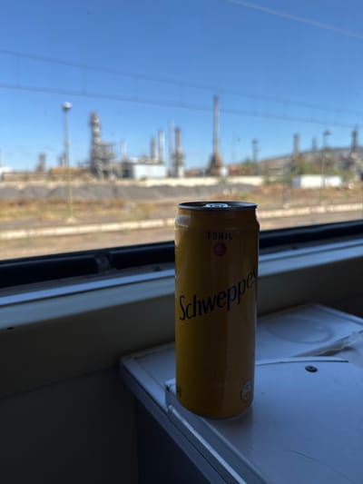 A yellow aluminum can of "Schweppes Tonic" stands upright on a white, possibly foldable, tray table inside a train compartment. Minor condensation is visible on the can's surface. Through the train window, an extensive industrial complex characteristic of an oil refinery is visible under a clear blue sky. This complex features numerous large metallic structures, including tall distillation towers, interconnected pipes, and storage tanks. The ground outside the train appears arid, with visible rail tracks immediately adjacent. The scene is brightly lit by natural daylight. No individuals are present. This industrial landscape, viewed from a train, is located in Sidi Kacem, Morocco, consistent with the SAMIR refinery.