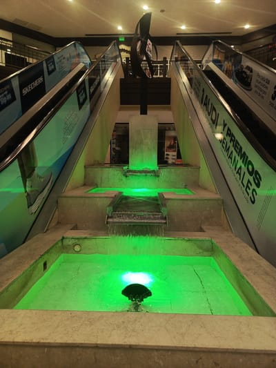 This indoor photograph, taken in Samborondón, Ecuador, showcases a modern commercial interior, likely a shopping mall. The central focus is a multi-tiered water fountain illuminated with bright green LED lights, creating a striking contrast with the surrounding neutral-toned concrete structure. Water gracefully cascades between the tiers into various pools.

Flanking the fountain are two parallel escalators ascending to an upper floor. Both escalators feature large, vibrant advertisements on their side panels. The left escalator displays "SKECHERS" branding with phrases like "Slip-ins" and "HANDS FREE," along with Spanish text such as "SIN TOCAR" (without touching). The right escalator advertises a "3 AUDI Y PREMIOS SEMANALES" (3 AUDI AND WEEKLY PRIZES) promotion, with details reading "Promoción válida del 20 de abril al 19 de julio" (Promotion valid from April 20 to July 19) and "Por cada $25 acumulables participa por uno de los" (For every $25 accumulated, participate for one of the...). A "SALIDA" (Exit) sign is visible near the top of the right escalator.

Above the escalators, an abstract, dark sculpture or decorative element stands prominently. The background reveals portions of upper levels with railings and ceiling lighting. The scene is brightly lit by artificial light, suggesting a commercial space that operates under controlled lighting conditions. There are no people visible, allowing the architectural features and the illuminated water display to dominate the composition.