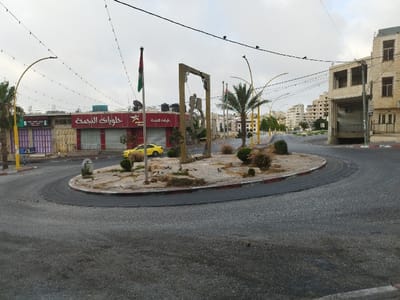 This image presents an elevated view of a traffic roundabout in the city of Dura, Palestinian Territory, under an overcast sky. The central, oval-shaped median is sparsely vegetated with low bushes and dry ground cover, featuring a small palm tree. A flagpole prominently displays a Palestinian flag (horizontal stripes of black, white, and green, with a red triangle at the hoist). Adjacent to the flag is a large, light-colored archway structure with intricate carvings or relief work. Several pigeons are visible on the median's surface.
The asphalt roadway surrounding the median appears damp in sections, with red and white painted curbs delineating its edges. Overhead, numerous string lights are strung across the intersection, connecting to yellow, curved street lamp poles; several small birds are perched on these wires.
Surrounding the roundabout are multiple urban structures. To the left, a single-story commercial building with a red facade features a large sign in Arabic, "حلويات النجمة" (Halawiyat Al-Najma), translated as "Star Sweets" or "Al-Najma Sweet," alongside a smaller English translation. Its windows are secured by closed metal shutters, and black water tanks are visible on its roof. A yellow sedan is parked or stationary near this building, partially obscured by the flagpole. To the right, a multi-story building constructed of light-colored stone exhibits multiple windows and a large, dark, ground-level opening consistent with a garage or loading bay, adjacent to a smaller arched entrance. In the background, numerous multi-story buildings of similar construction recede into the distance along the streets, characterizing an urban landscape. No human subjects are visible in the scene.