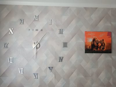 A large, modern wall clock with Roman numerals is the main subject of this image. The clock face itself is not visible, but silver Roman numeral markers are attached to the wall at their respective positions. The numbers visible are: X, XI, XII, I, II, III, IV, V, VI, VII, VIII, and IX. The word "TIME" is also spelled out in silver letters on the wall. The clock has two silver hands, one short and one long, indicating the current time. To the right of the clock, there is a framed piece of art depicting two lions, possibly a mother and cub, with a sunset in the background. The art is colorful, with shades of orange and red dominating. The wall itself has a textured geometric pattern of light and dark grey diamond shapes. The image appears to be taken indoors, likely in a living room or hallway, in Lisakovsk, Kazakhstan. There is a white ceiling molding visible at the top of the image. The overall impression is one of contemporary decor.