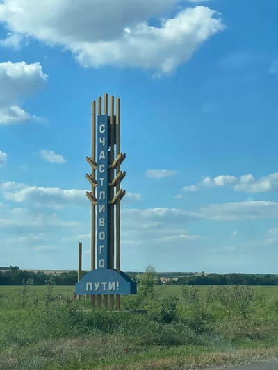 A vertical, blue and white sign with Cyrillic lettering stands in a grassy area under a partly cloudy blue sky. The sign is framed by vertical wooden posts and has a semicircular blue base. The top portion of the sign reads "СЧАСТЛИВОГО" vertically, and the base reads "ПУТИ!" horizontally. The surrounding environment consists of green grass, some wildflowers, and in the background, a treeline and fields under a bright, sunny sky with scattered white clouds. The location is identified as the city of Donskoe, Russia.