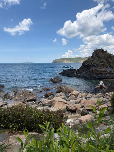 The image depicts a rocky coastline under a blue sky with scattered white clouds. The sea is a deep blue, with small waves lapping against the shore. The shoreline is covered in various sizes of rocks and boulders, with some green vegetation growing between them and in the foreground.
In the mid-ground, a cluster of dark, layered rocks juts out into the sea, forming a small promontory. There are a few small boats anchored in the water near this rock formation. Two people, dressed in light-colored clothing and hats, are visible sitting on the rocks on the right side of the image, seemingly looking out at the sea. A rope barrier with stanchions and a sign are present on the dark rock formation.
The horizon shows a distant island or peninsula with hills under the sky. The overall impression is of a scenic, natural coastal landscape, likely a tourist spot. The location is identified as Phú Lương, Vietnam.