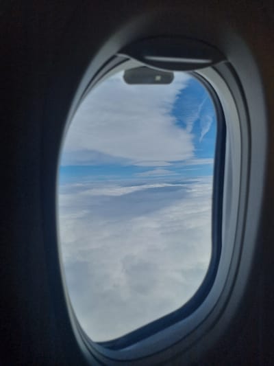 The image shows a view from inside an airplane, looking out of a window. The window is oval-shaped with a gray frame and a sliding shade partially open. Outside, there are clouds and a strip of blue sky visible. There is no indication of the location being Brakyovtsi, Bulgaria in the image. The focus is on the view from the airplane window. No people or specific objects besides the airplane window and clouds are visible.