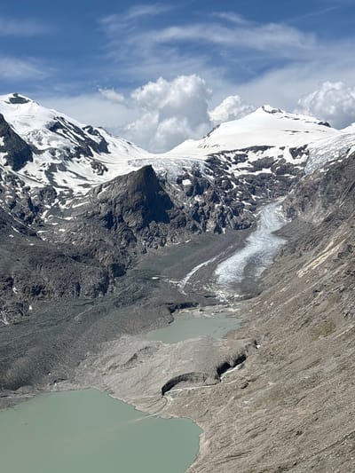 The image depicts a high-altitude glacial landscape. In the foreground, a pale green lake fills the lower portion of the frame. The lake is bordered by a rocky, debris-strewn shore that rises to form steep, gray mountain slopes. Further up the slopes, a prominent glacier, characterized by its white ice and dark crevasses, snakes its way down from the higher peaks. The upper reaches of the image are dominated by snow-capped mountains with stark, dark rock formations exposed in places. The sky above is a bright blue, dotted with fluffy white clouds.

The scene is identified as being in Heiligenblut am Großglockner, Austria. The image captures the Pasterze Glacier and its proglacial lake, likely taken from a viewpoint on the Glocknerstraße (Grossglockner High Alpine Road). There are no people or animals visible in the image. The environment is rugged and natural, indicative of a mountain wilderness.