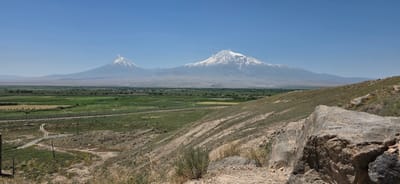 The image depicts a scenic view of Mount Ararat, a dormant volcano, in the distance, seen from a hilly, grassy terrain in the foreground. The mountain, capped with snow, dominates the horizon under a clear blue sky. In the mid-ground, a fertile valley with agricultural fields and scattered trees stretches towards the mountain. A road or path is visible winding through the fields. The foreground consists of a rocky, sloped hillside with sparse vegetation and large stones. The overall impression is one of natural grandeur and a peaceful rural landscape. This view is characteristic of the area around Lusarat, Armenia, known for its proximity to Mount Ararat. There are no people or animals visible in the frame. The lighting suggests it is a sunny day.