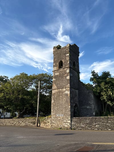 The image shows a stone tower, which appears to be part of an old ruin, standing against a bright blue sky with wispy clouds. The tower is tall and rectangular, made of rough-hewn stones, with arched openings visible at the top. Green vegetation is growing on the tower and along the top of the stone wall in front of it. Trees with lush green foliage surround the tower on the left and right. In the foreground, there is an asphalt road with yellow lines. A wooden pole stands to the left of the tower, and a stone wall borders the road. A sign attached to the tower reads "PARISH OF CHUIRCE THEAGHTA ABBEY of the HOLY CROSS". This indicates the location is the Abbey of the Holy Cross in Cahersiveen, Ireland. There are no people or animals visible in the image. The lighting suggests it is daytime, likely late afternoon given the long shadows.