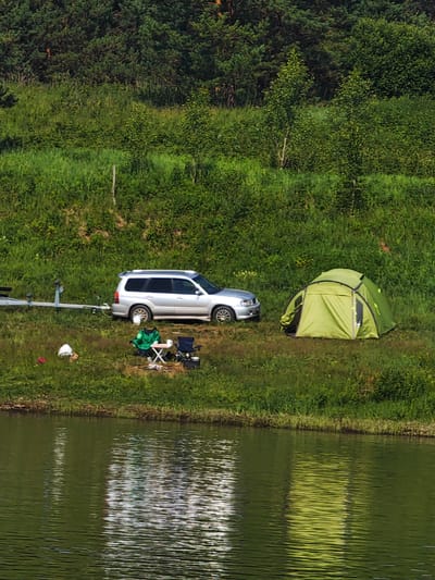 The image depicts a campsite on the grassy bank of a body of water, likely a river or lake, with a dense forest in the background. A silver SUV is parked on the grass, hitched to a boat trailer. A green tent is pitched nearby. In front of the SUV, a person is seated in a green jacket at a small folding table with two chairs, suggesting a moment of rest or activity. Several white plastic bags and a red object are scattered on the grass. The water in the foreground reflects the trees and sky, creating a textured surface. The overall scene suggests an outdoor recreational activity.