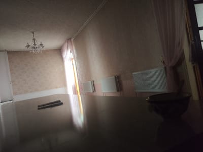 A dimly lit room with a chandelier hangs from the ceiling. The walls are covered in patterned wallpaper, and there are radiators along one wall. A large dark table dominates the foreground, reflecting the light from a window on the left. On the table, two cylindrical objects are placed side-by-side, and a dark bowl with a spoon rests to the right. A sheer pink curtain is visible on the right side of the room, near a dark doorframe. The scene appears to be an interior of a room in Shahimardanabad, Uzbekistan.