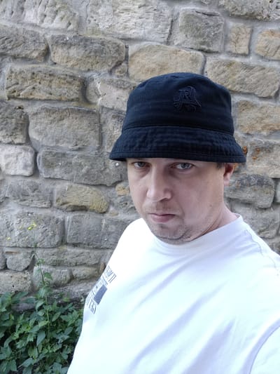 A man wearing a black bucket hat with an embroidered emblem and a white t-shirt stands in front of a stone wall. The man is positioned on the right side of the frame, facing forward with his head slightly tilted. The stone wall, constructed of large, rough-hewn blocks of varying shades of grey and beige, occupies the background. To the lower left of the frame, a small cluster of green plants with a single yellow flower is visible. The lighting suggests it is daytime. The image appears to have been taken in Doksy, Czechia.